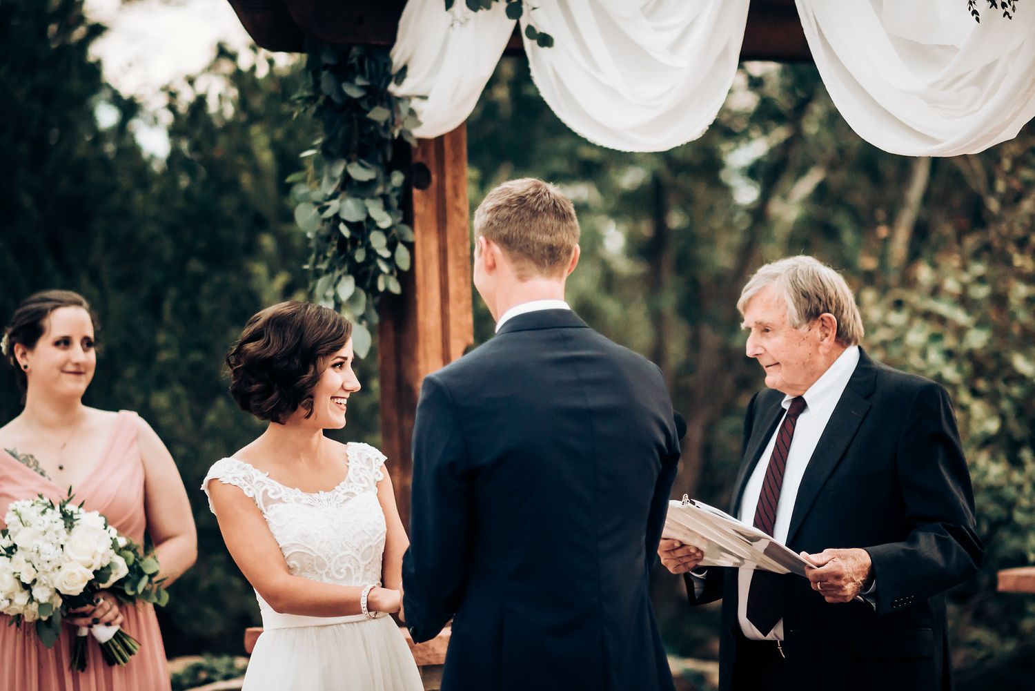 Outdoor wedding ceremony under a white draped pergola with stone pillars and forest backdrop.