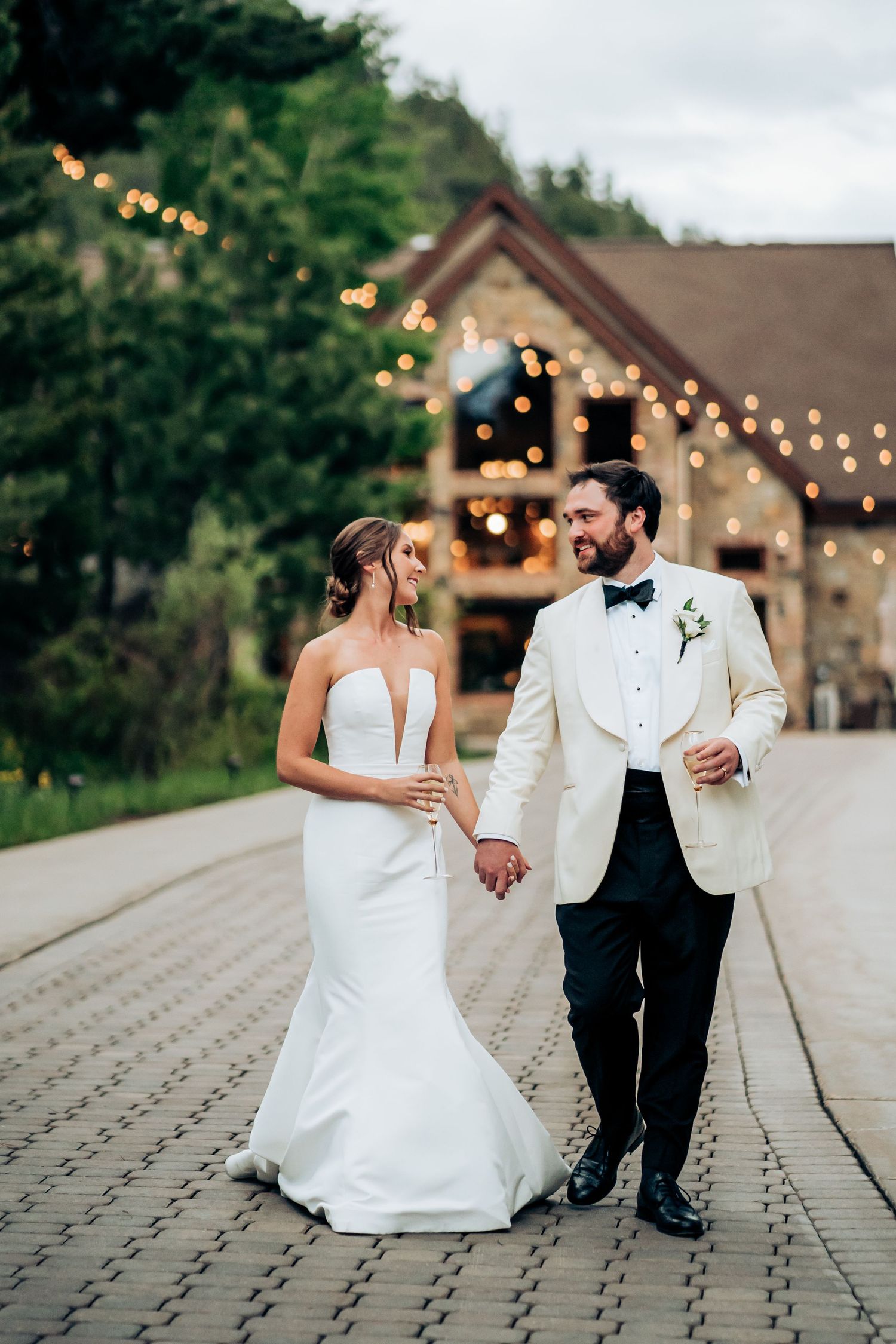 Elegant couple walking hand in hand outside rustic venue with twinkling lights.