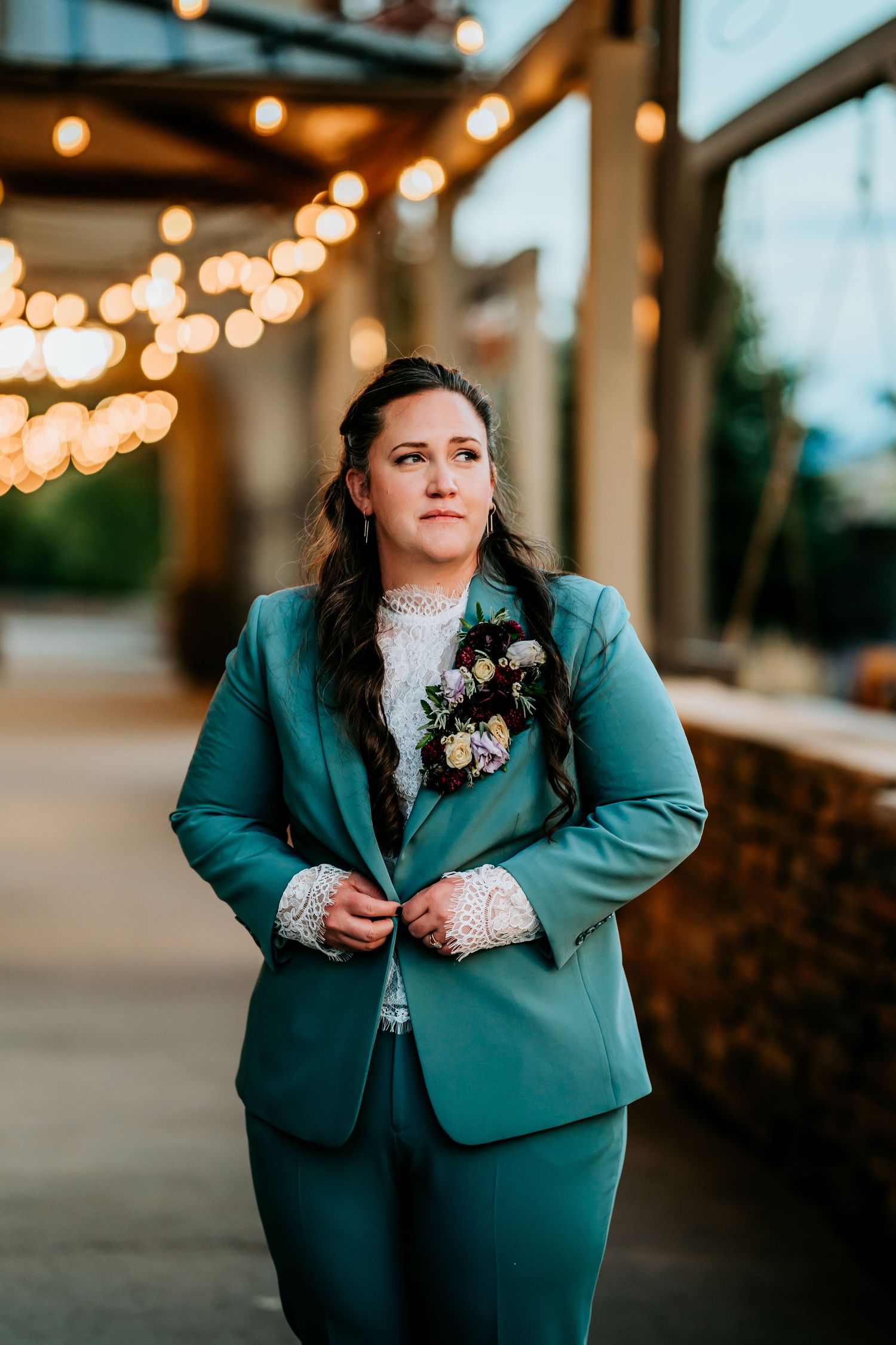 Person in teal wedding suit adjusting jacket with floral boutonniere under warm string lights.
