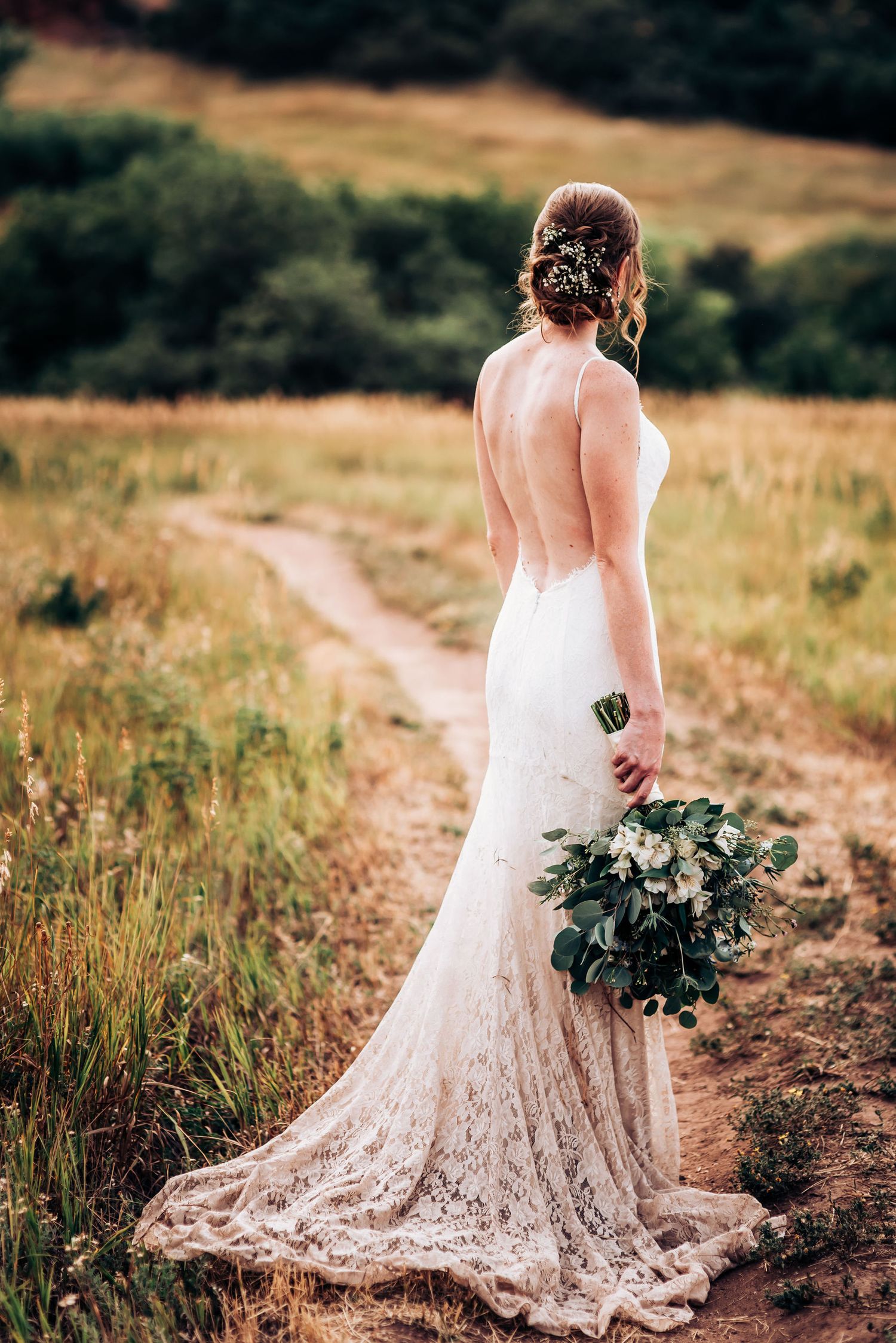 A bride in a backless lace gown holds a bouquet while standing on a mountain trail.