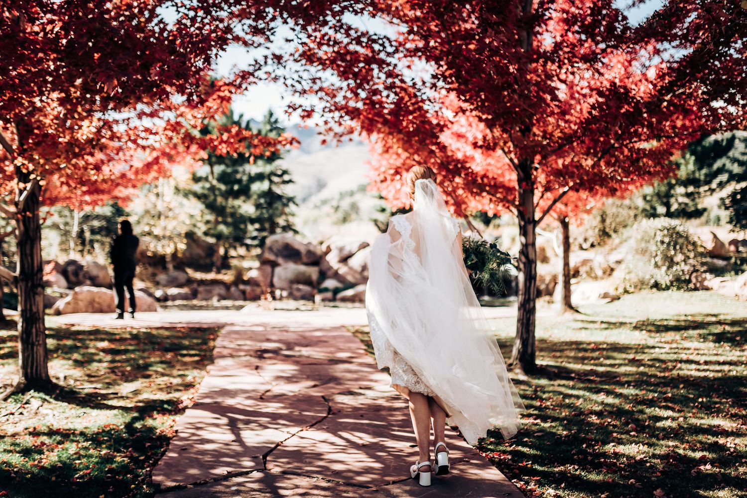 Bride in flowing dress walking under vibrant red autumn trees on a garden path.