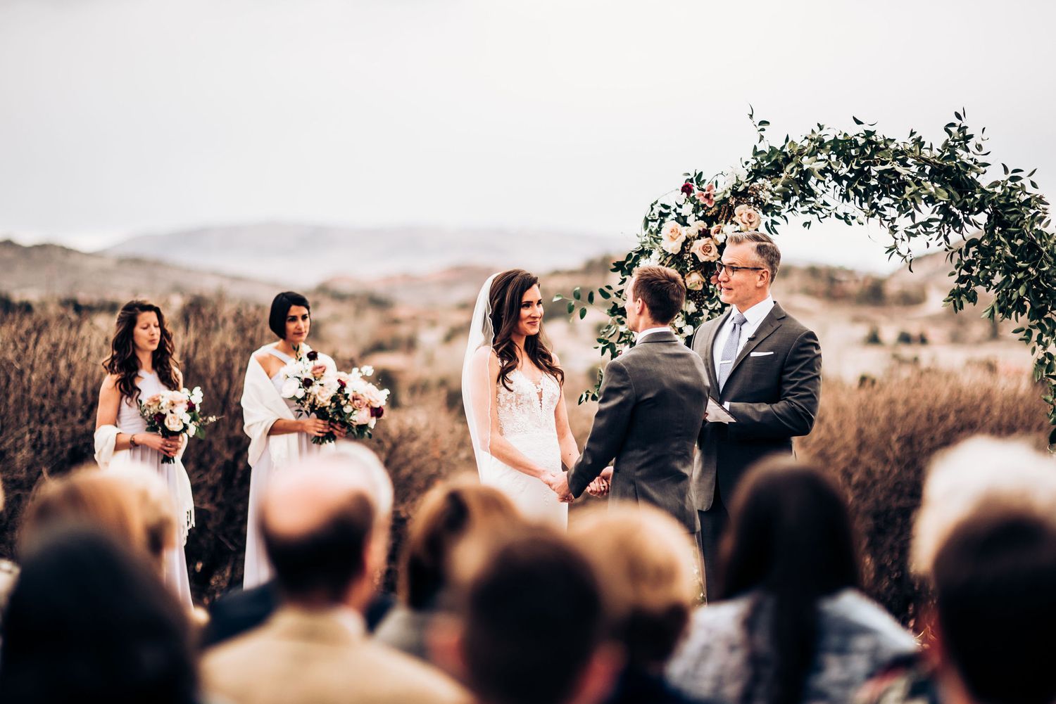 An intimate outdoor wedding ceremony with guests watching beneath a floral arch in a scenic landscape.
