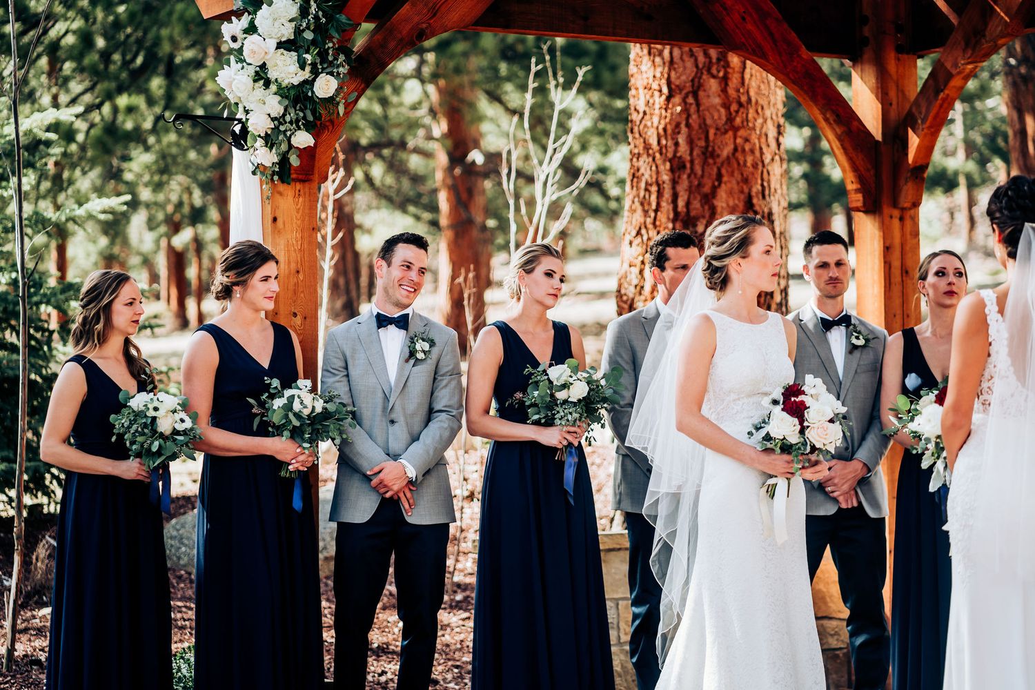 Wedding party in navy dresses and gray suits standing under wooden arbor during ceremony.
