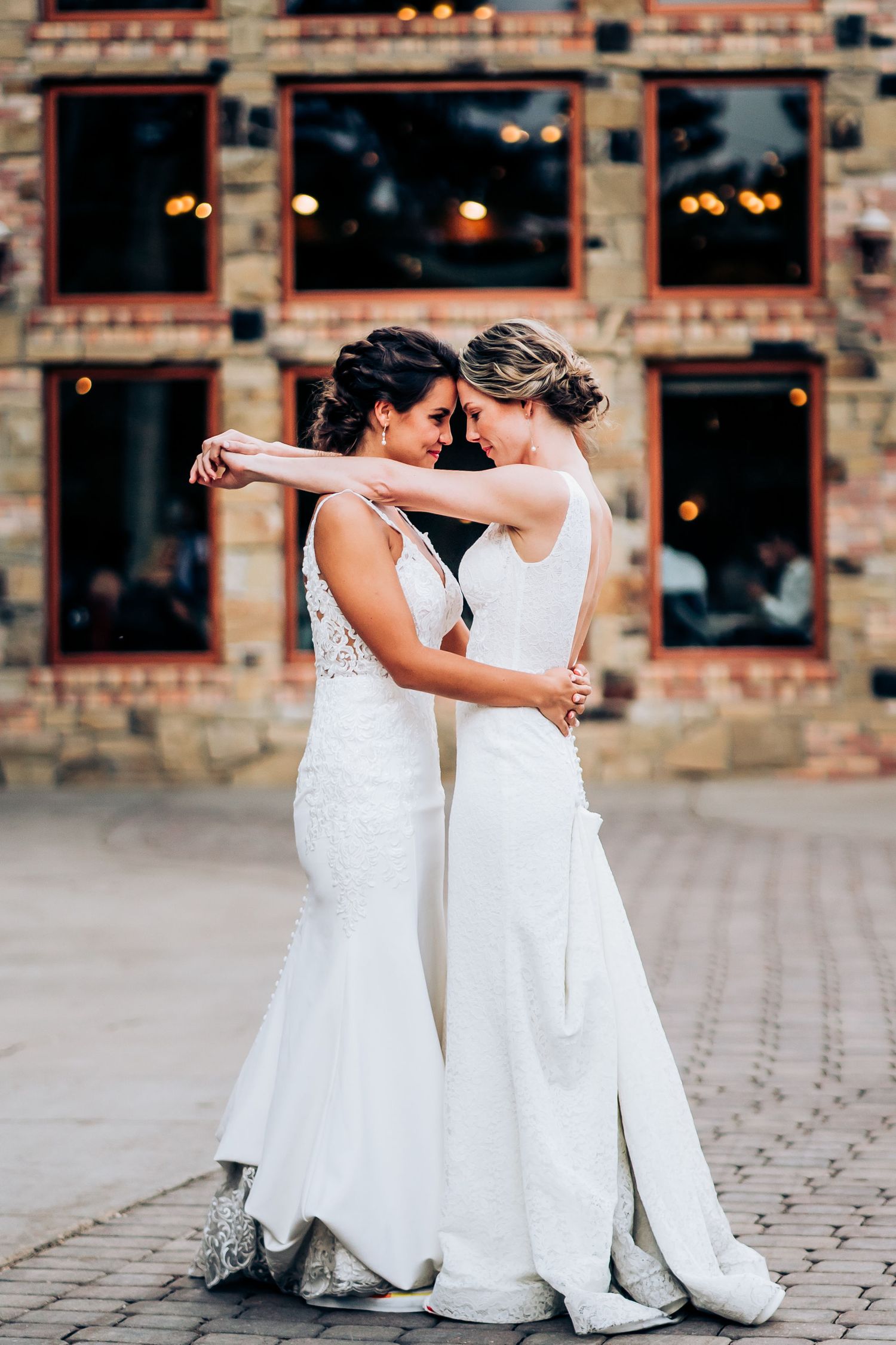 Two people in elegant white wedding gowns dance together in front of a rustic brick building with glowing windows.