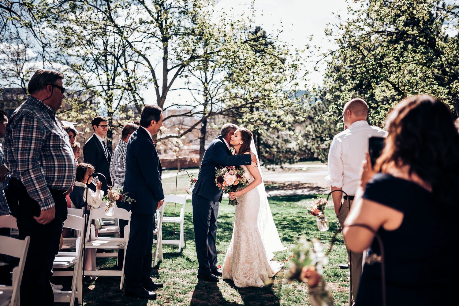 Outdoor wedding ceremony in progress with guests watching under blooming spring trees.