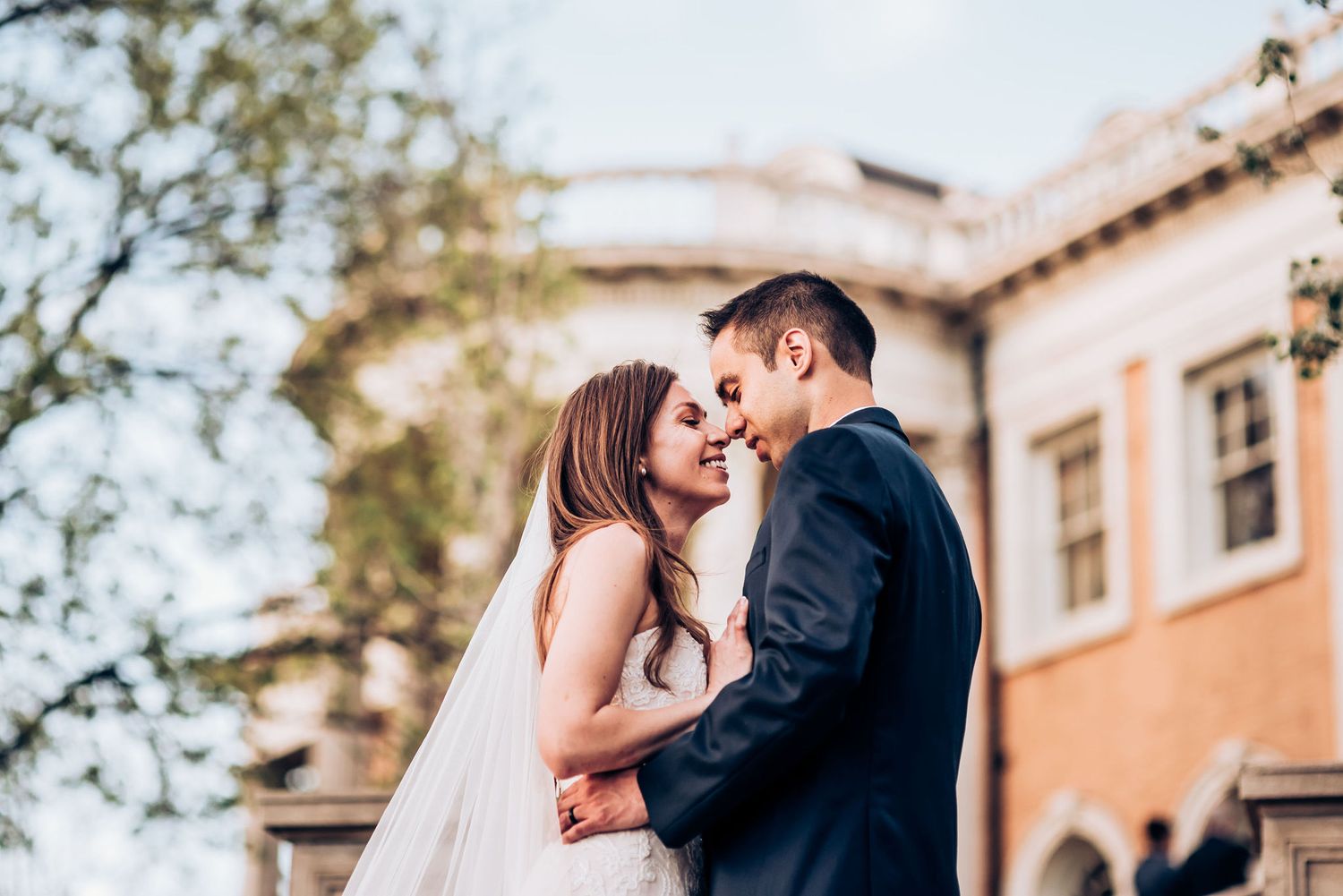 Romantic moment between newlyweds against peach-colored mansion exterior.