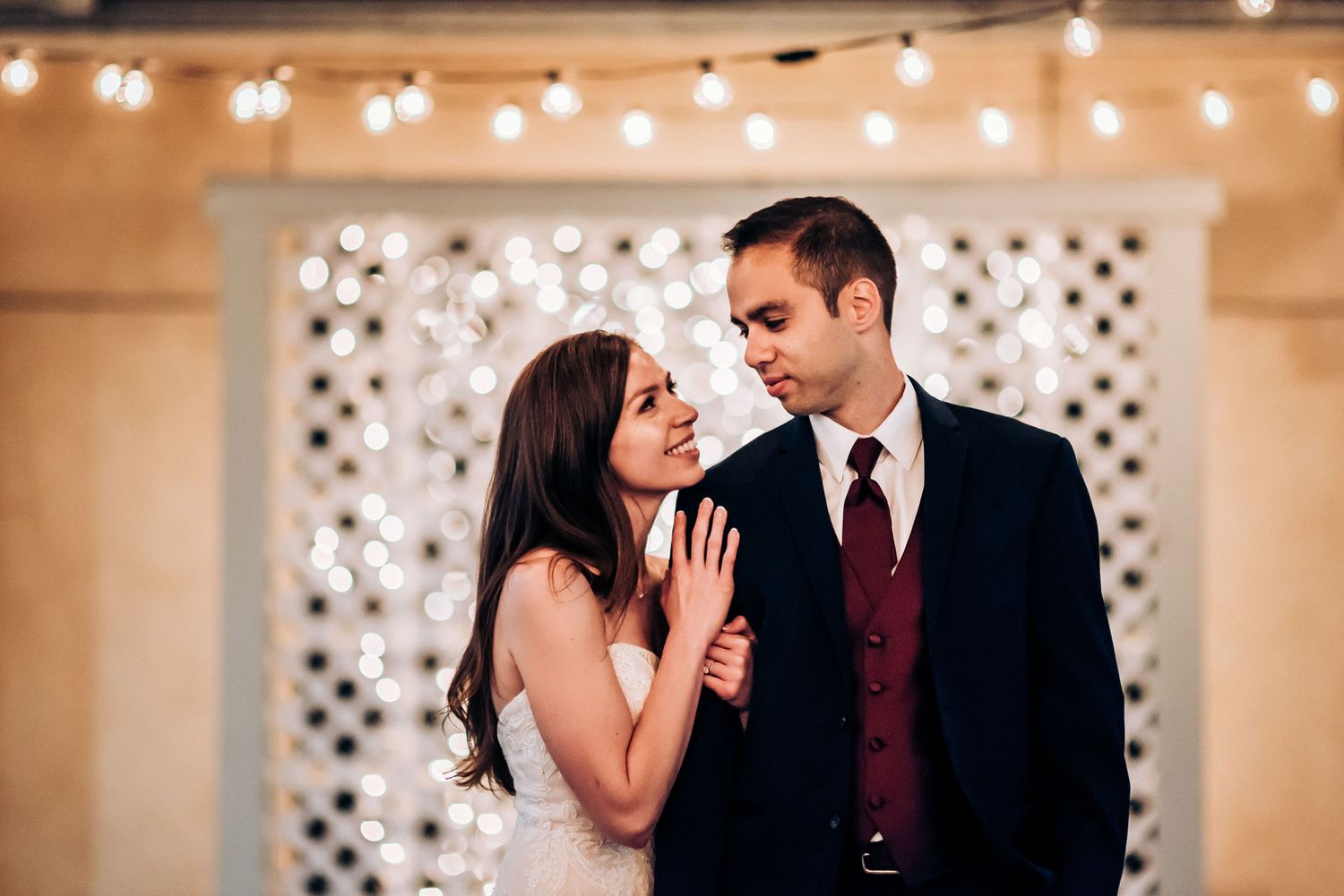 A couple shares a romantic moment against a backdrop of glowing fairy lights and decorative lattice.