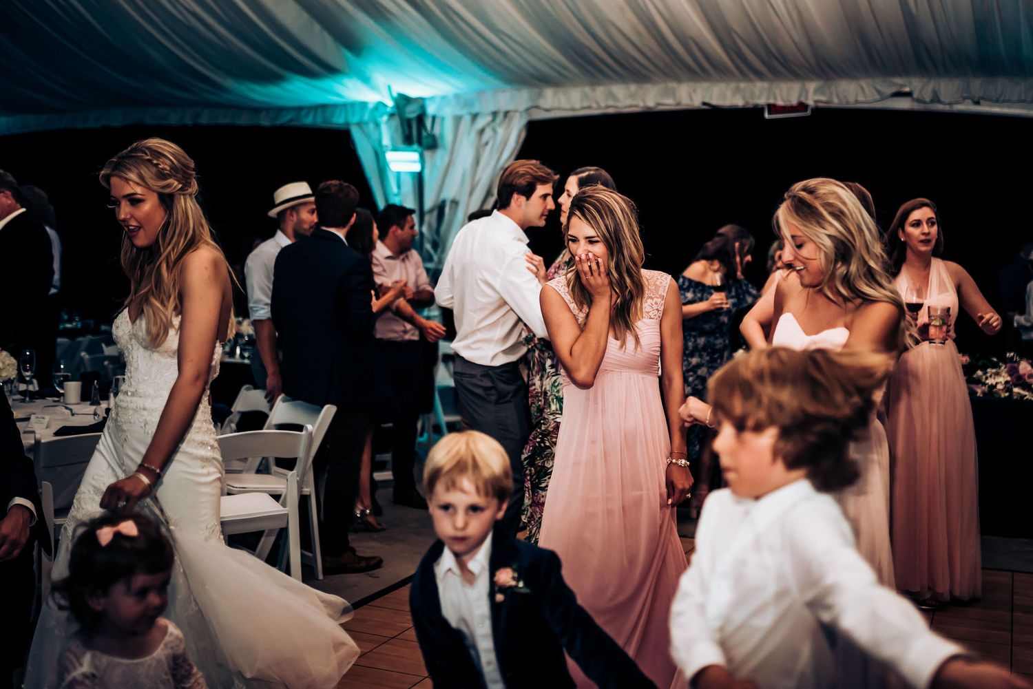 Wedding reception guests dance and celebrate under decorative tent lighting.