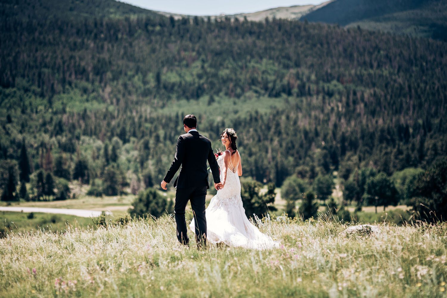Newlyweds walk hand in hand through mountain meadow with forest valley in background.