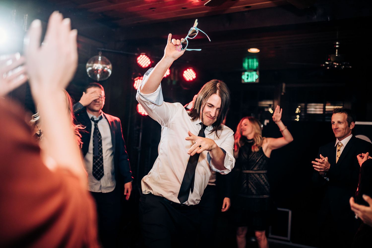 Wedding guests dance energetically at a dimly lit reception with hands in the air.