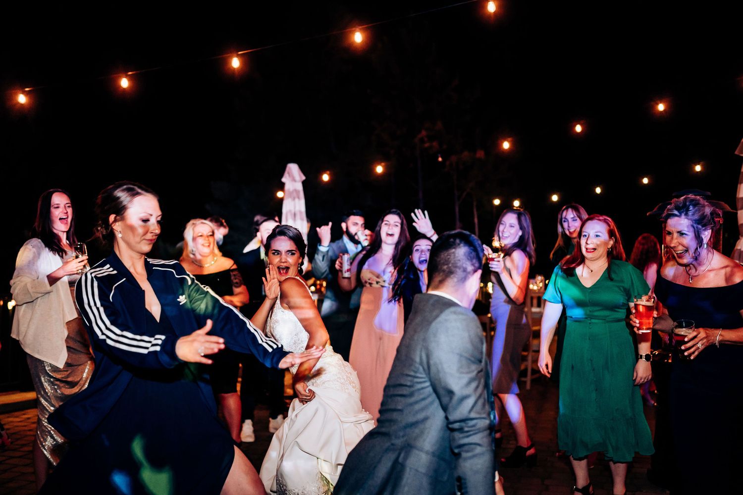 Energetic dance party at an outdoor wedding reception with string lights overhead.
