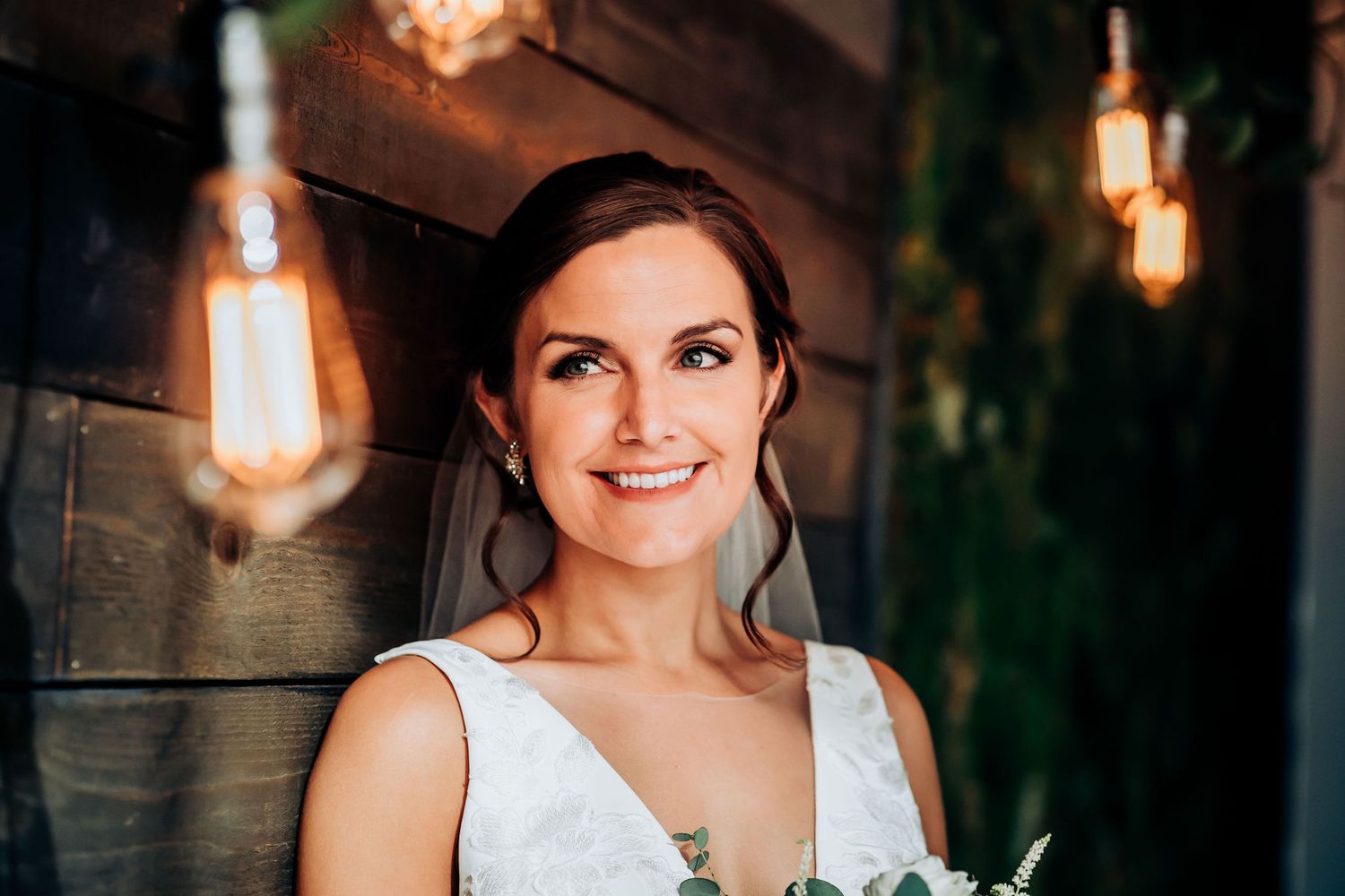 Bride in white V-neck dress smiling warmly under ambient lighting with greenery backdrop.