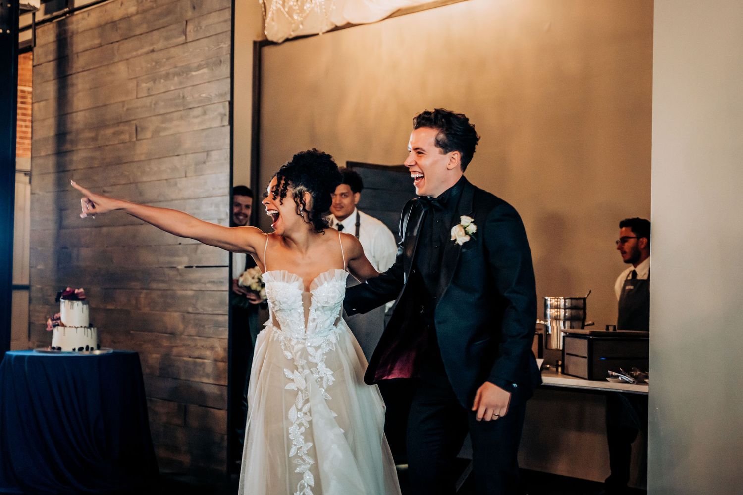 Wedding couple gesturing excitedly at reception venue with exposed brick walls.