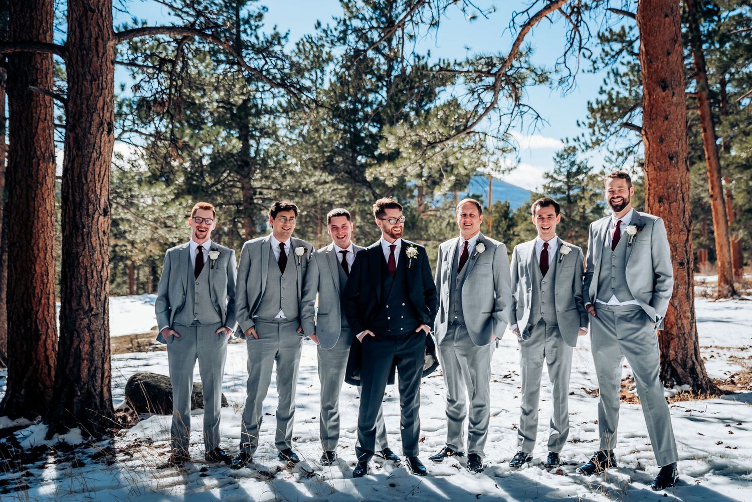Groomsmen in light gray suits pose with navy-suited companion in snowy mountain forest.