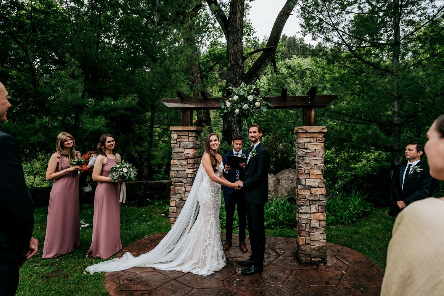 An intimate outdoor wedding ceremony with attendants in formal attire gathered under a decorated stone arch.