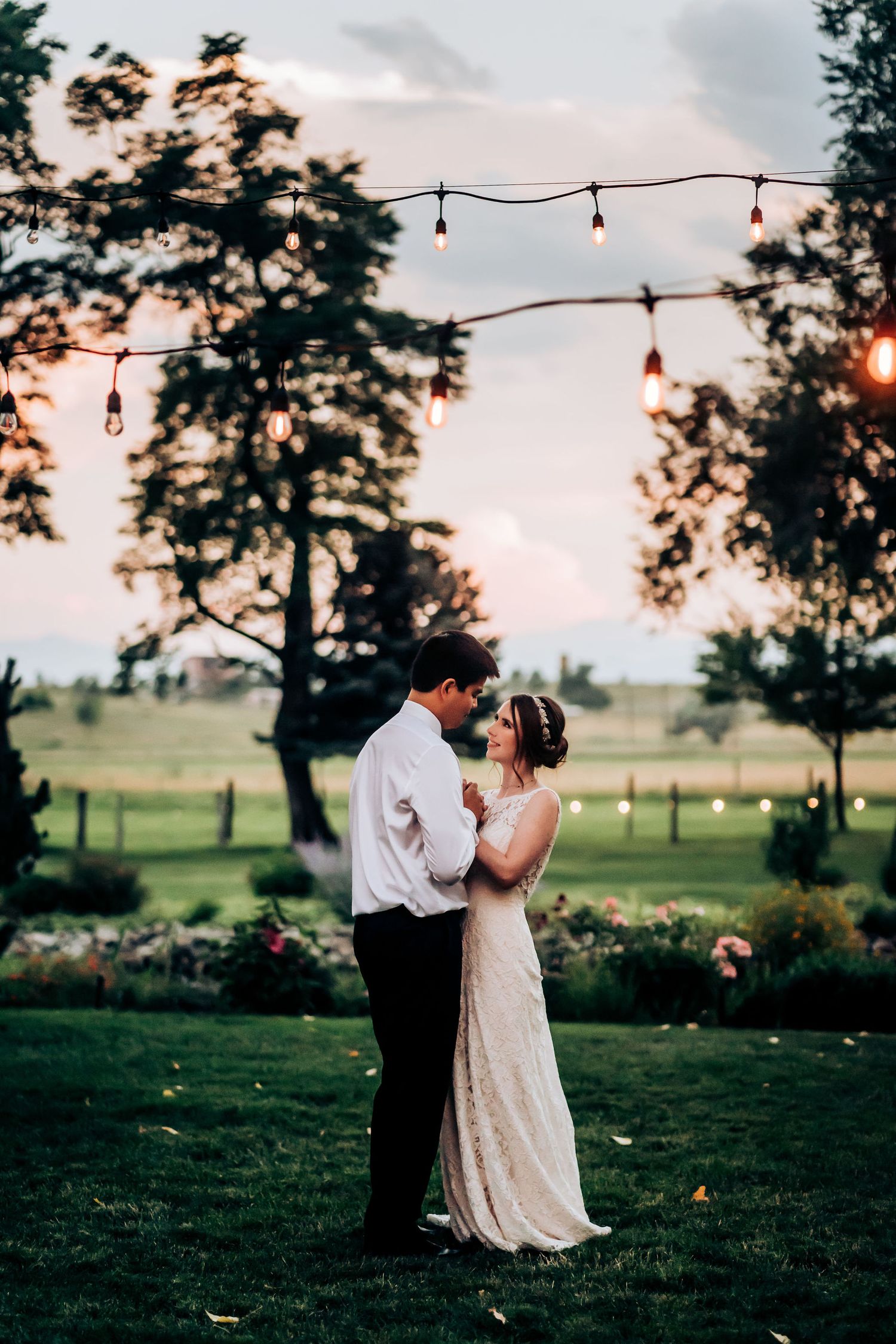 A couple shares a romantic dance on a garden lawn beneath string lights at sunset.
