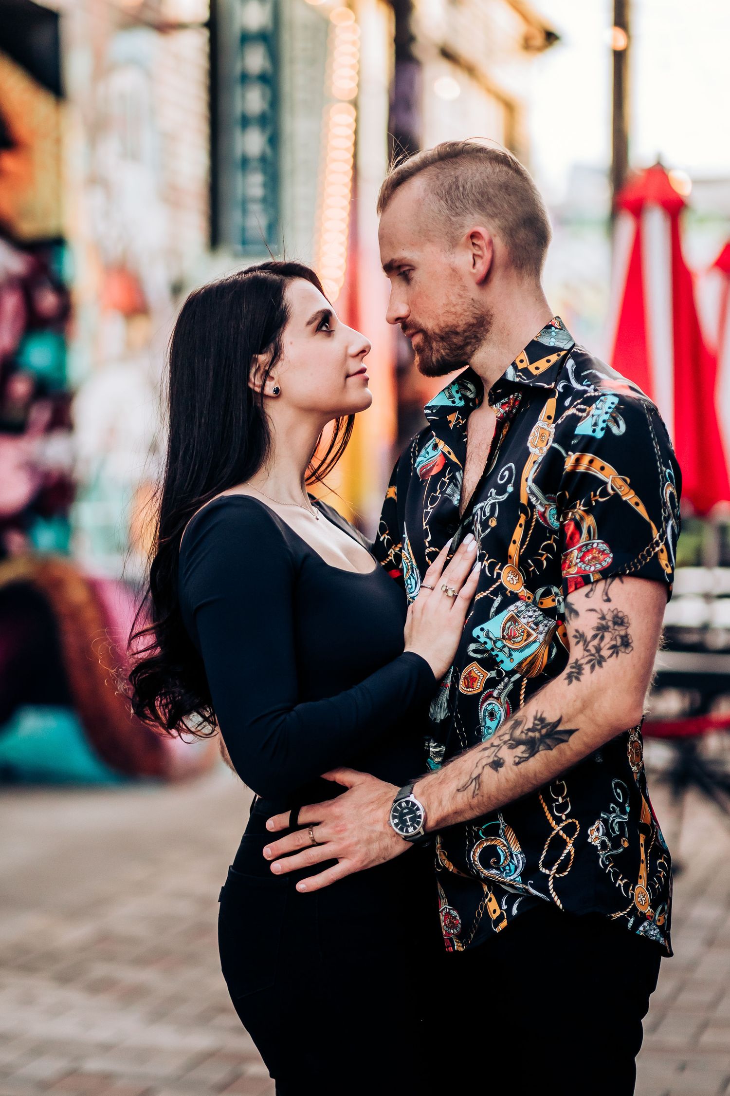 Romantic couple embraces against colorful urban backdrop with man wearing patterned shirt and woman in black.