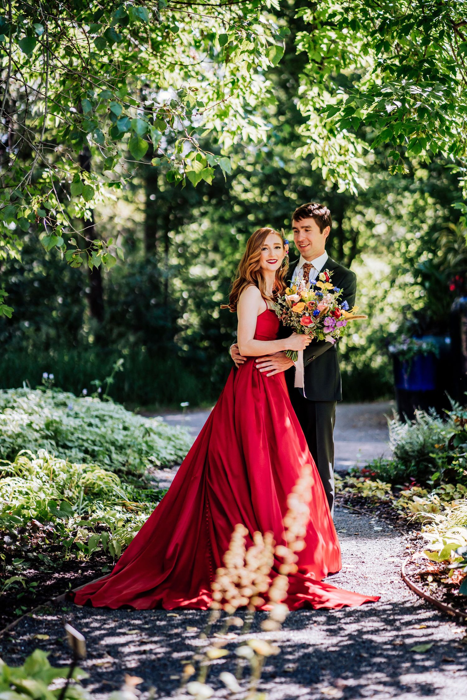 A romantic garden portrait featuring a red ball gown and formal attire amidst dappled sunlight.
