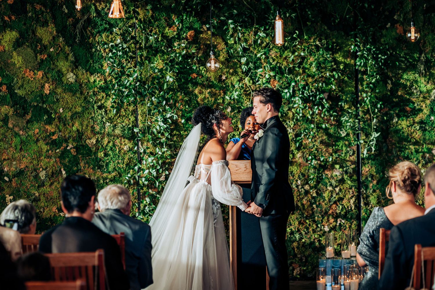 Wedding ceremony moment against dramatic green wall with hanging lights.
