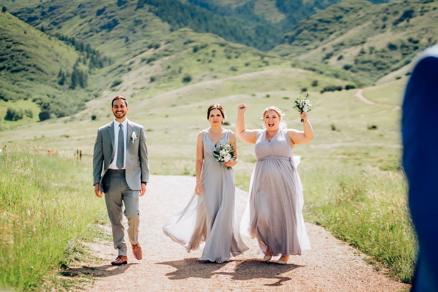 Wedding party members walking and celebrating on dirt path through mountain meadow.
