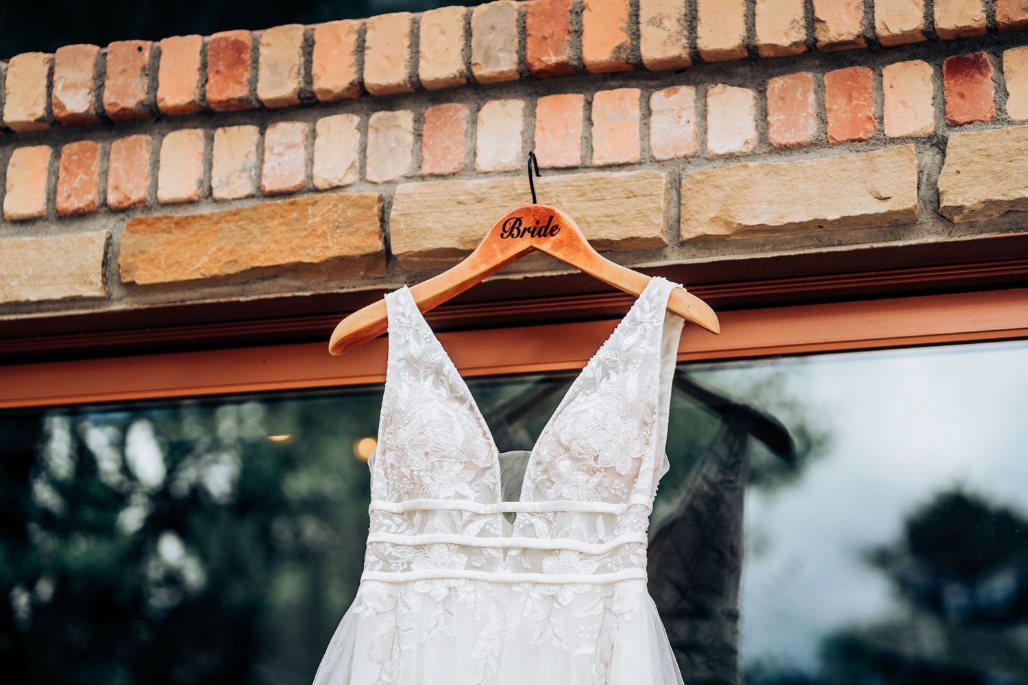 A white lace wedding dress hangs on a wooden hanger against a brick wall.