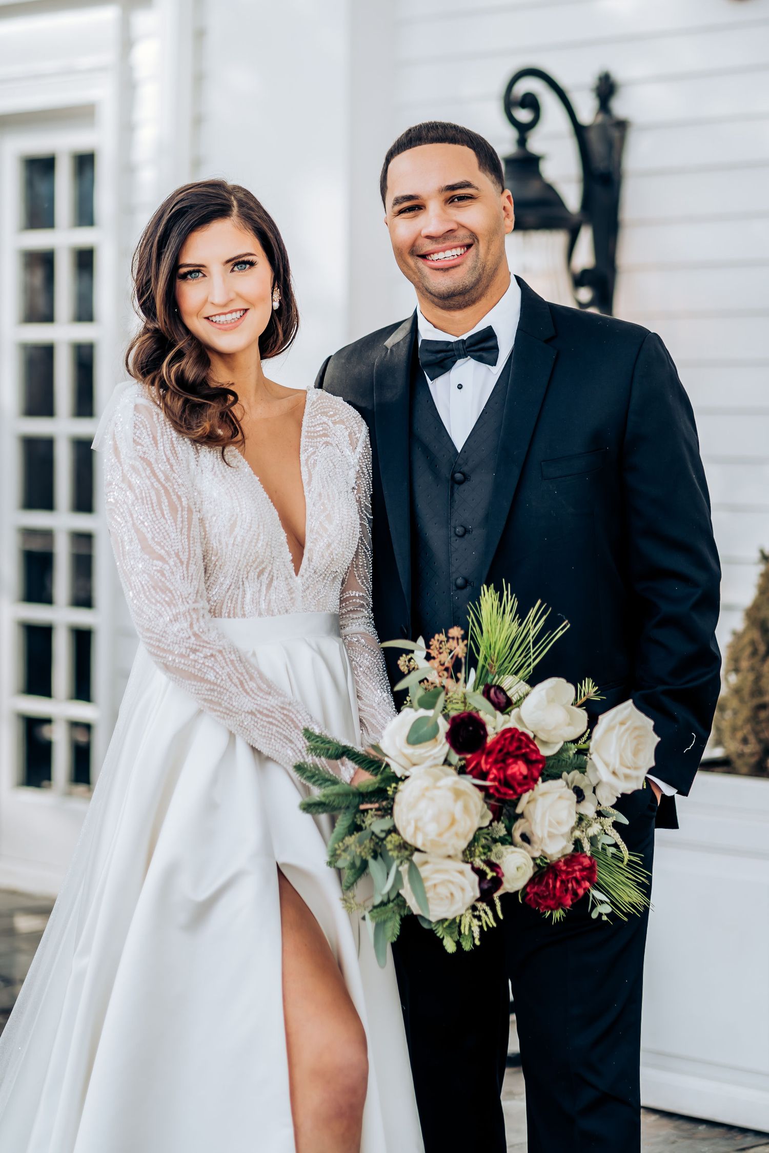 Elegant wedding couple in formal attire posing against white building with floral bouquet.