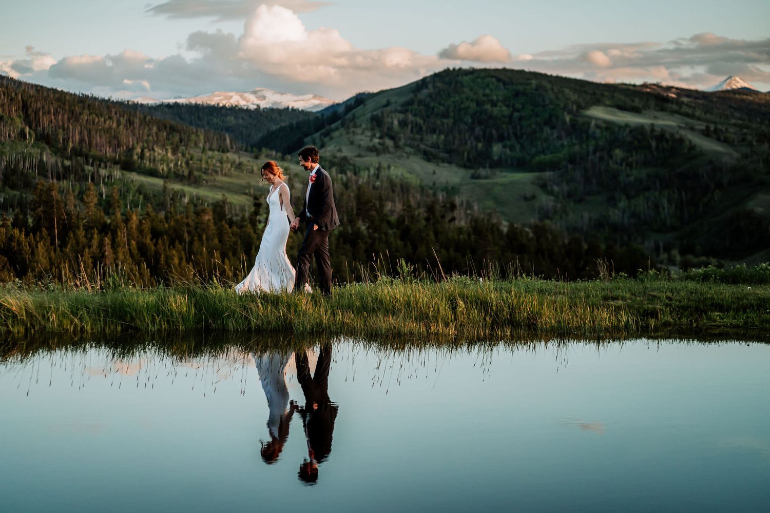 Newlyweds pose by serene mountain pond with mirror-like reflection at dusk.