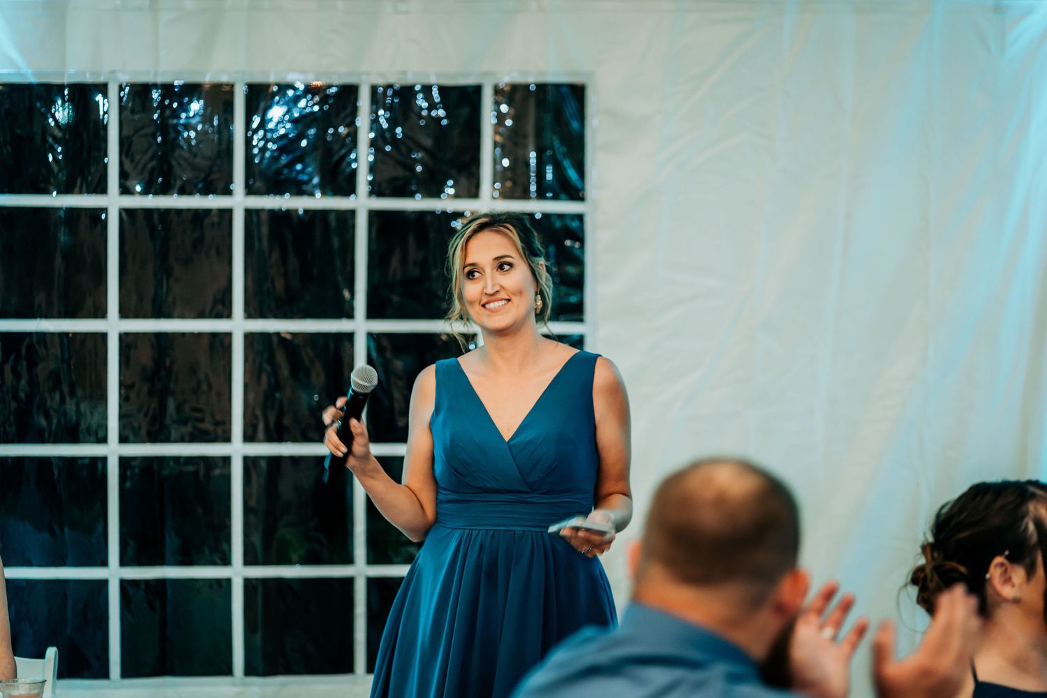 Wedding reception speech being given in front of large windows at evening venue.