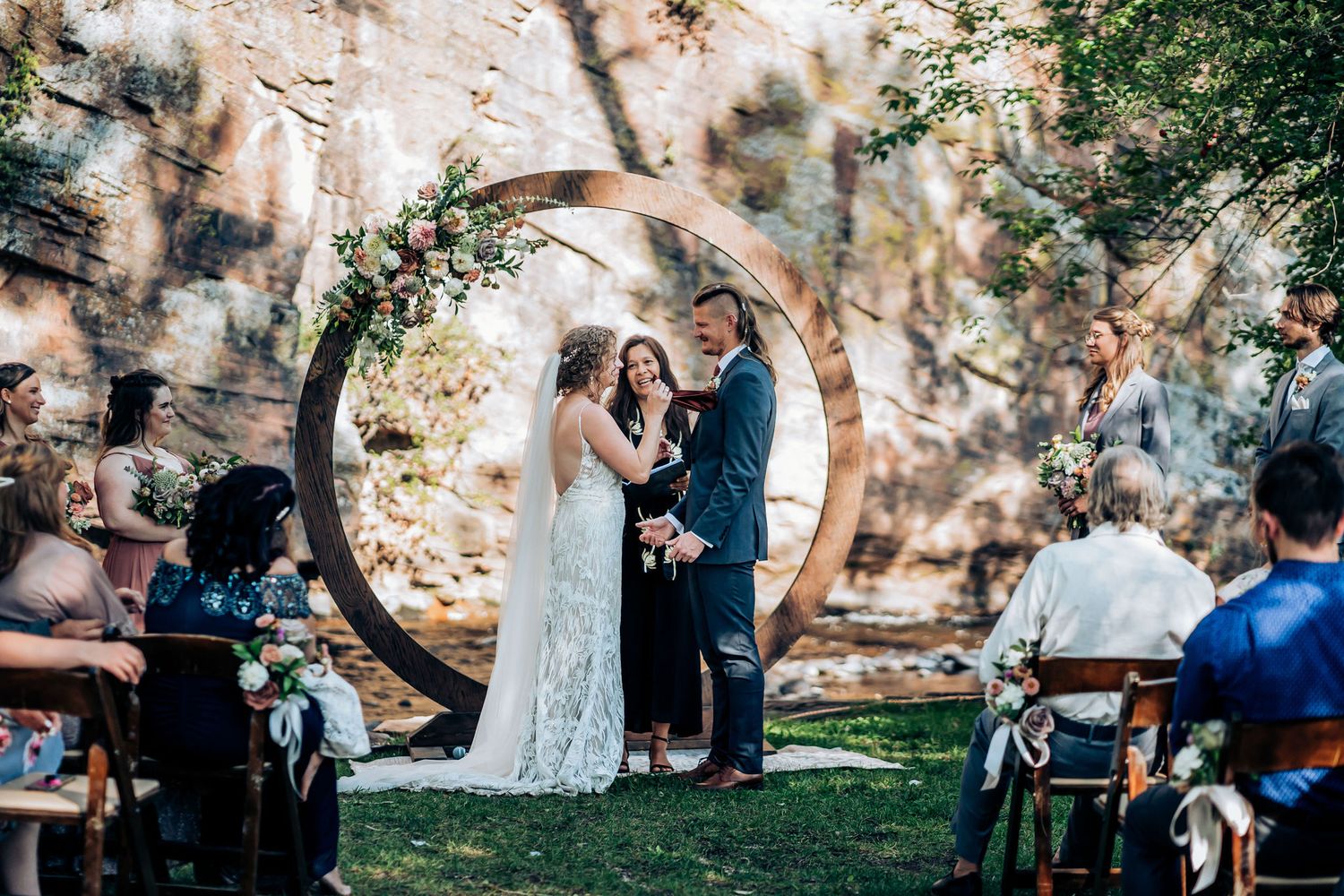 A wedding ceremony under a large circular arch decorated with flowers against a rocky backdrop.