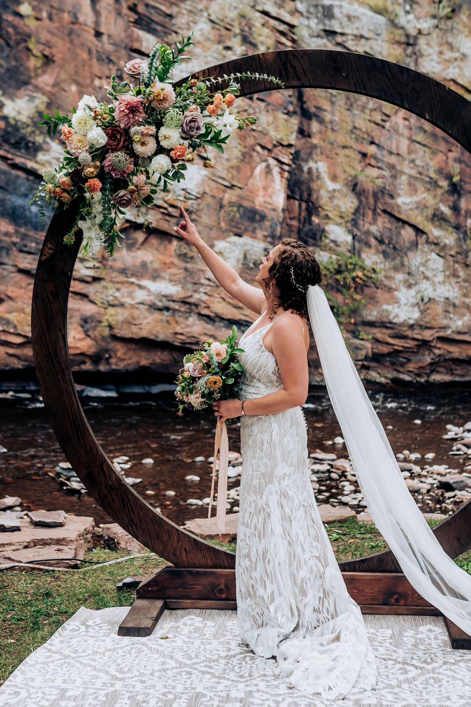 Bride arranges flowers on circular wedding arch against dramatic rock wall background.