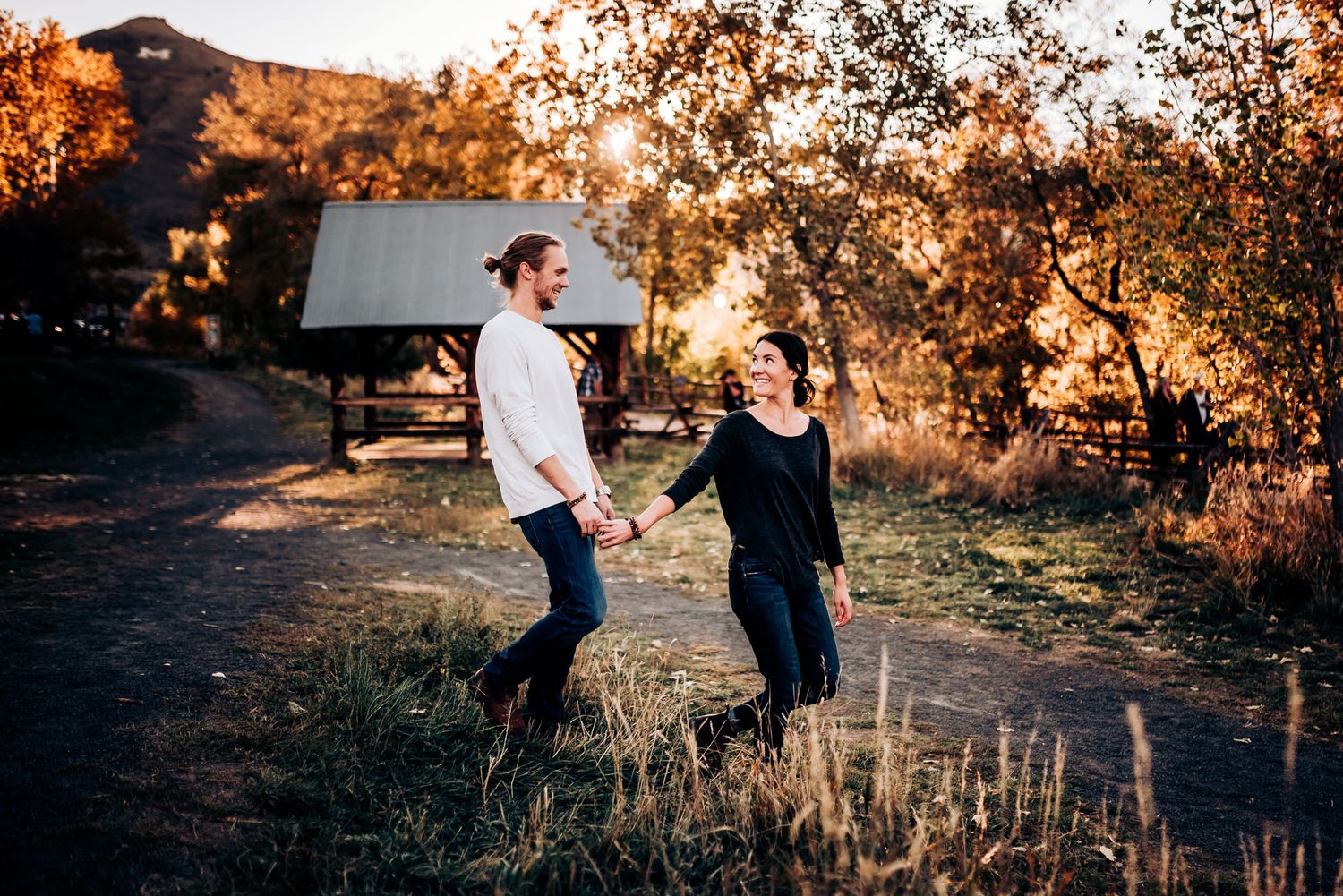 A couple stands together on a scenic mountain path at sunset near rustic buildings.