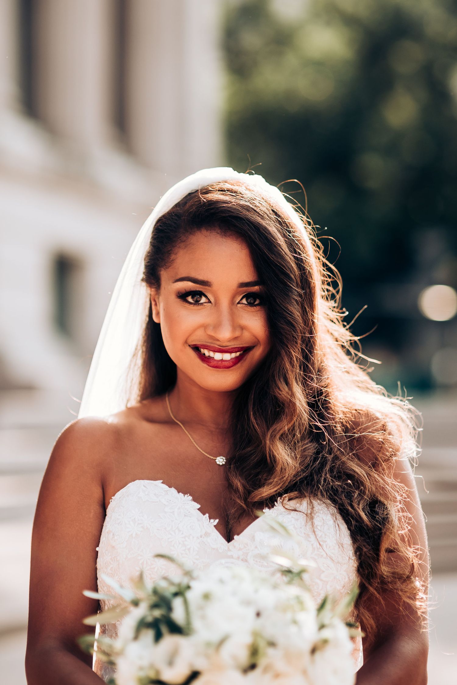 Bride in white strapless gown poses with bouquet against architectural backdrop.