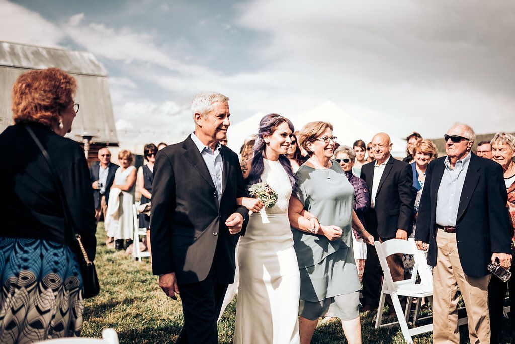 Wedding party processes down outdoor aisle with guests seated on white chairs.