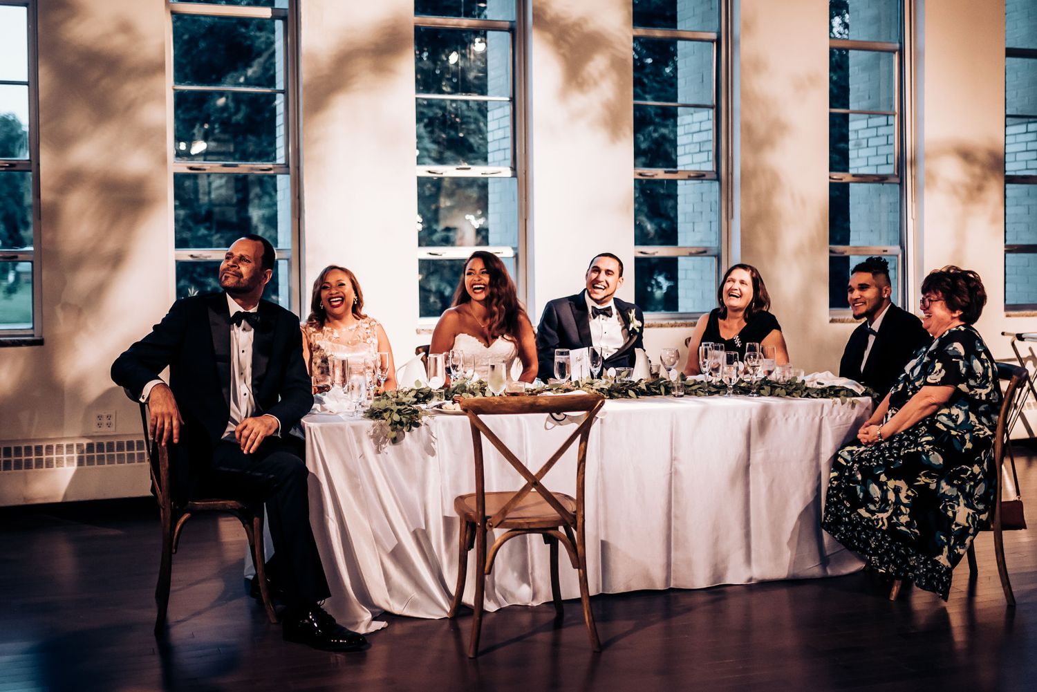 Formal wedding party seated at elegant head table with tall windows and soft lighting.