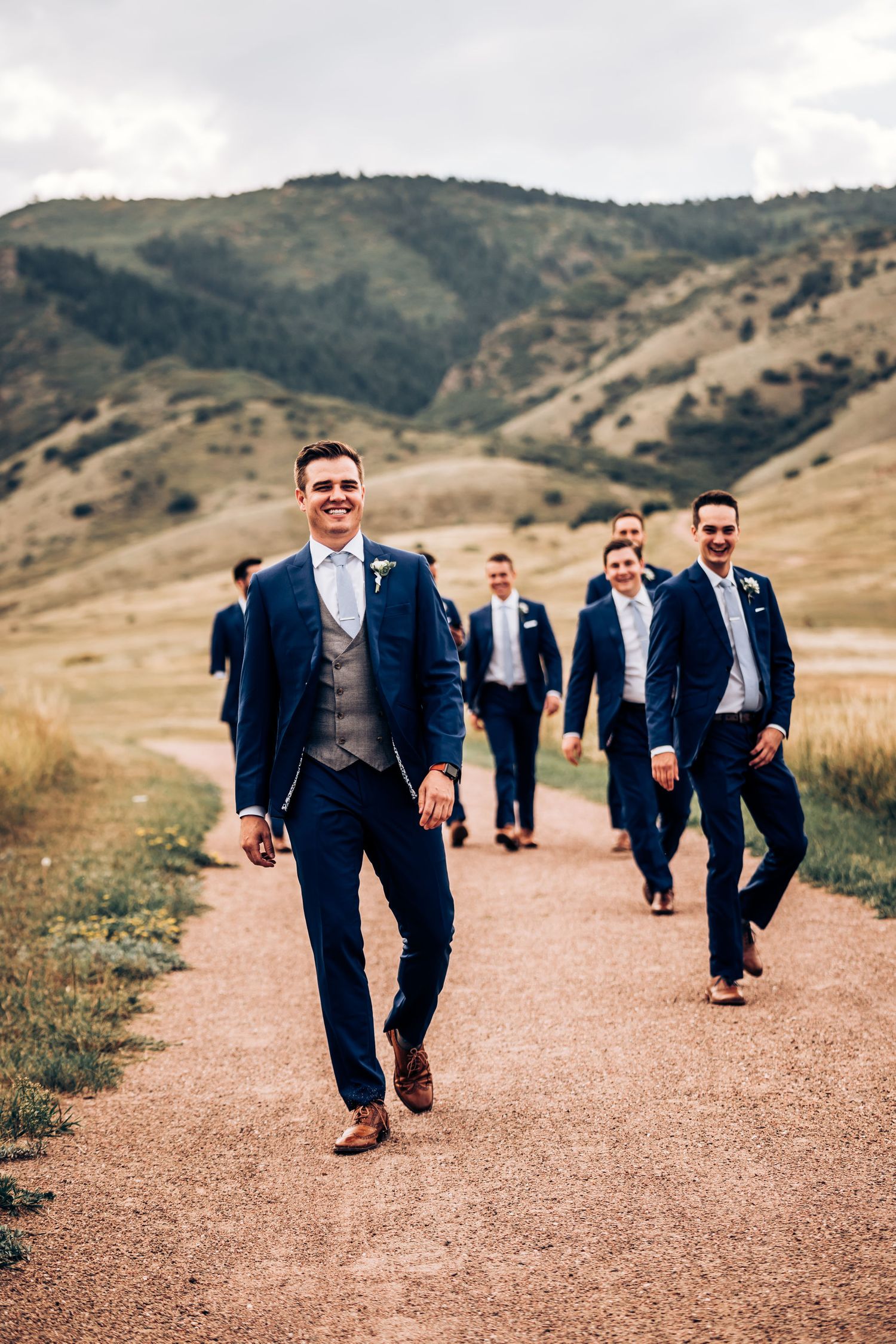 Groomsmen walking on mountain trail wearing navy suits against natural landscape.