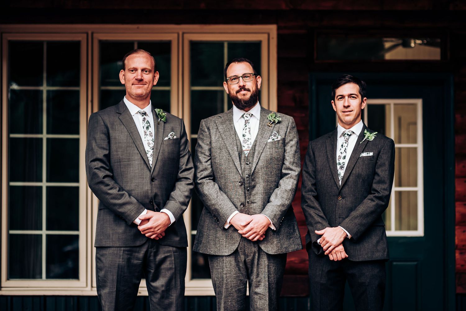 Three men in formal wedding attire pose together on a rustic porch.