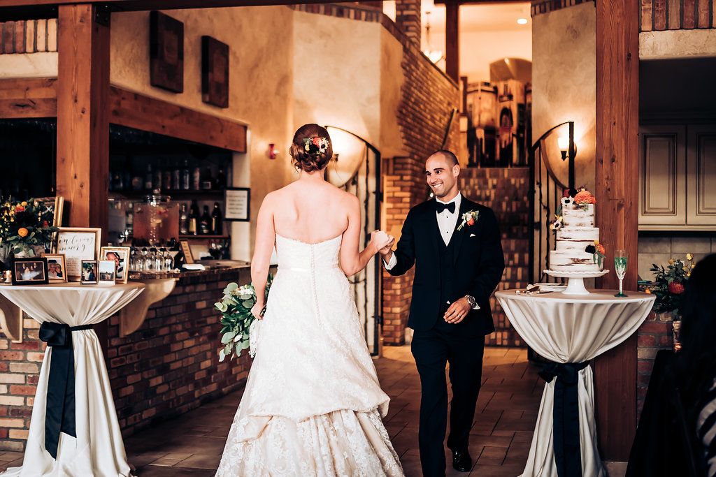 Newlyweds walk through rustic reception venue with tall cocktail tables and wedding cake.