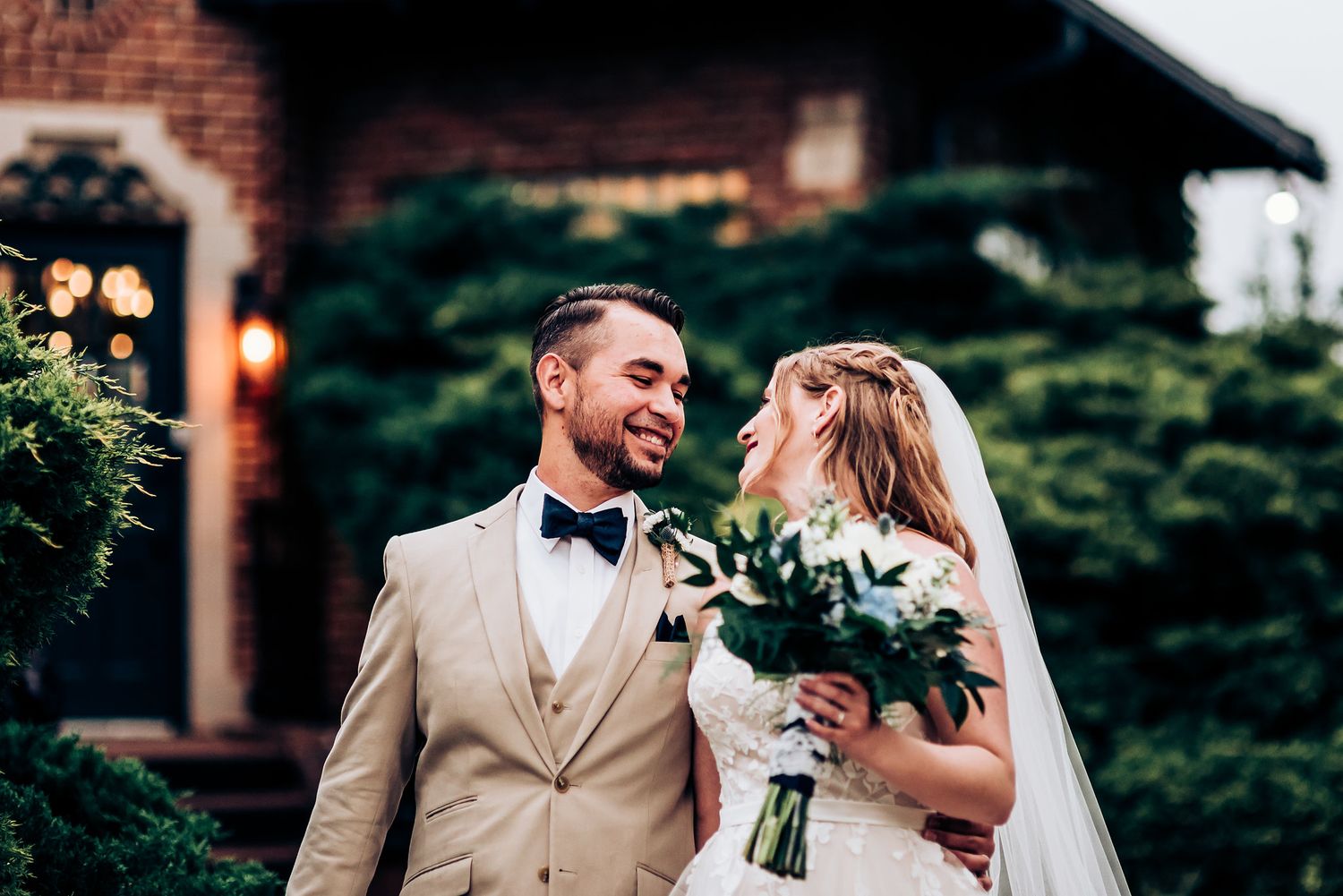 A romantic wedding couple shares a joyful moment in their elegant tan and white attire with a brick building backdrop.