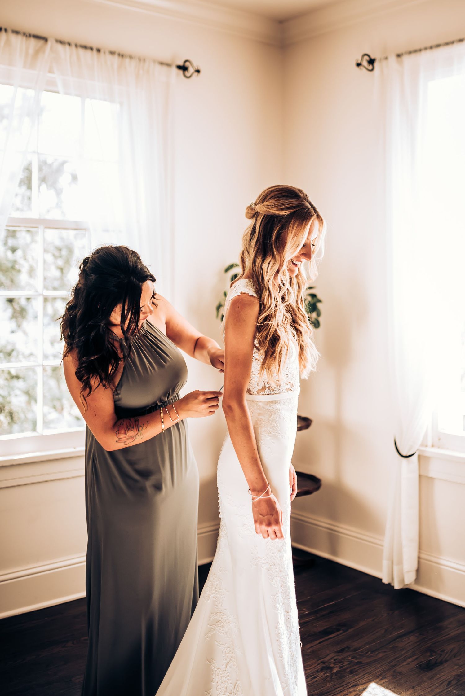 Bridesmaid in gray dress helps adjust wedding gown during bridal preparation.