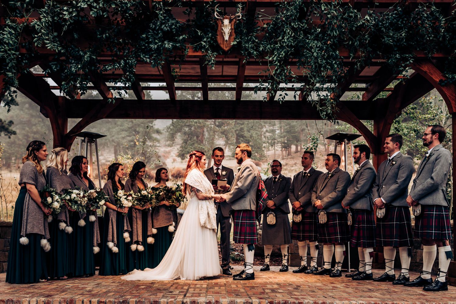 Wedding party lines up under pergola with bride in white dress and groomsmen in traditional Scottish kilts.
