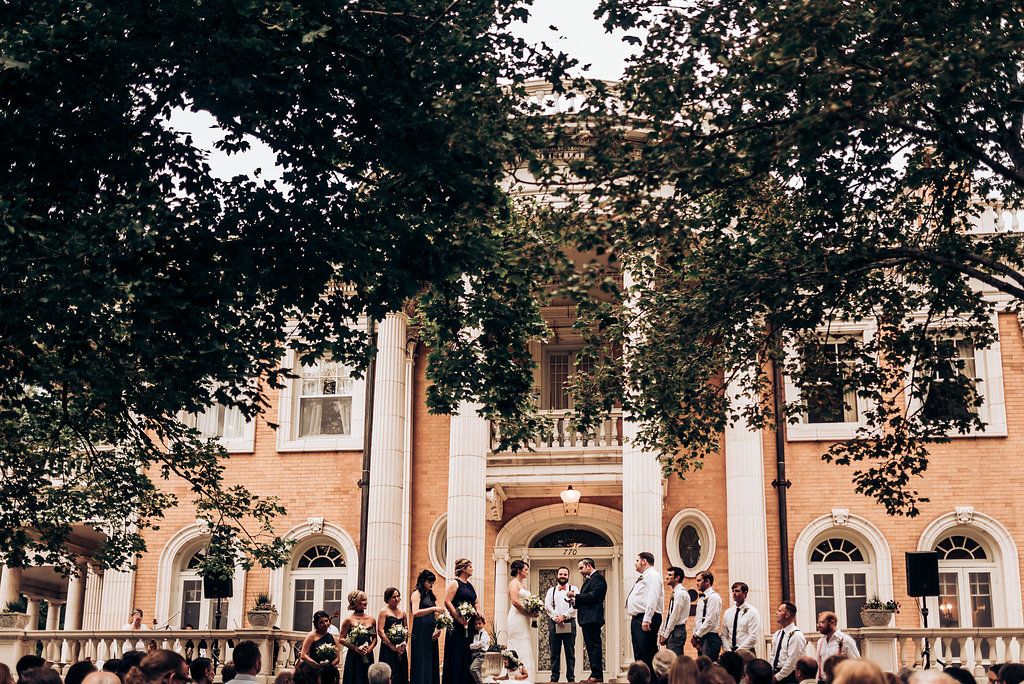 A wedding ceremony takes place on the steps of an elegant brick building with white columns and arched windows.