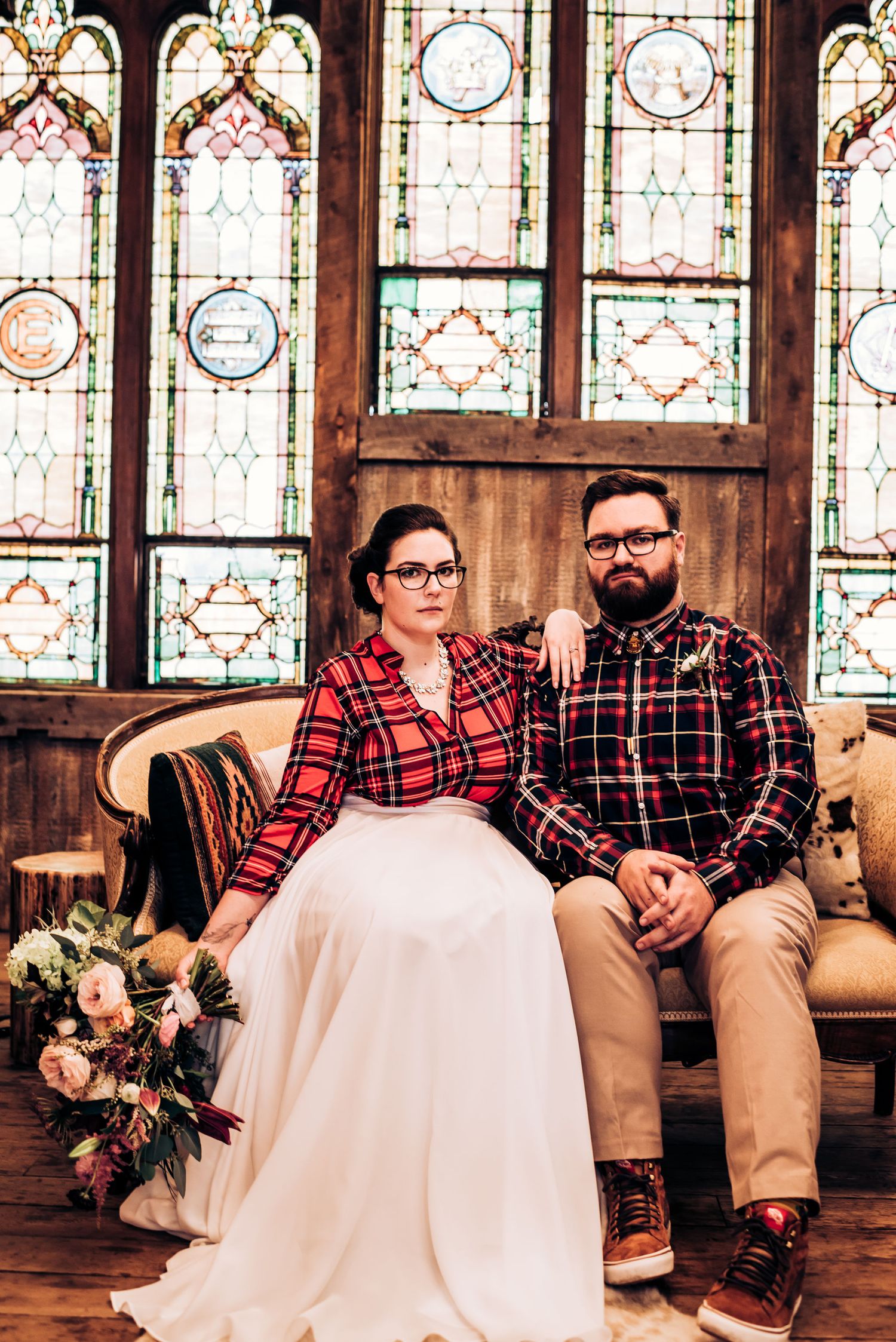 Couple in matching plaid shirts pose on vintage sofa against stained glass windows in rustic wedding setting.