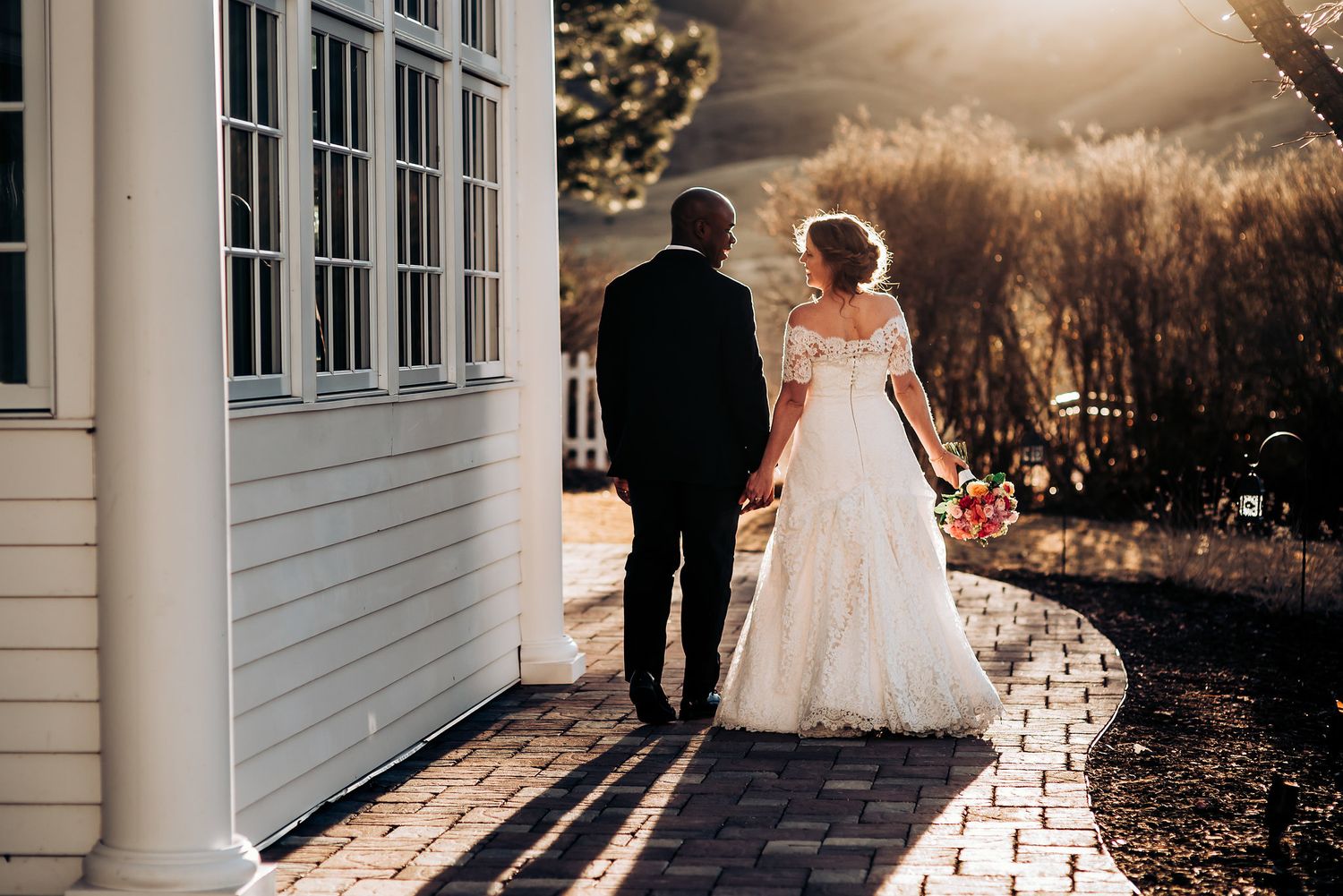 Wedding couple standing outside white building on curved brick pathway at sunset.