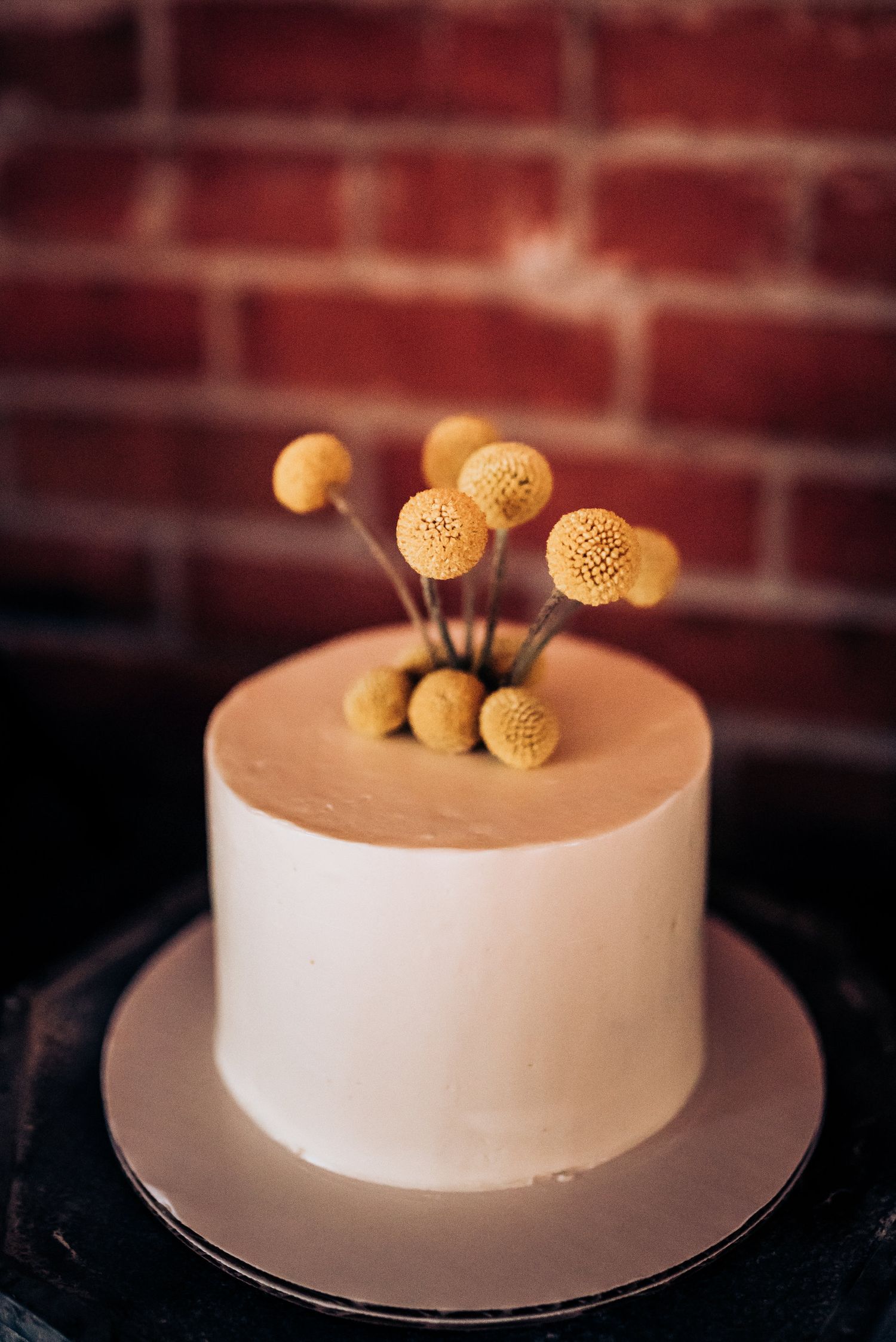 Simple white cake decorated with golden craspedia ball flowers against a brick wall backdrop.