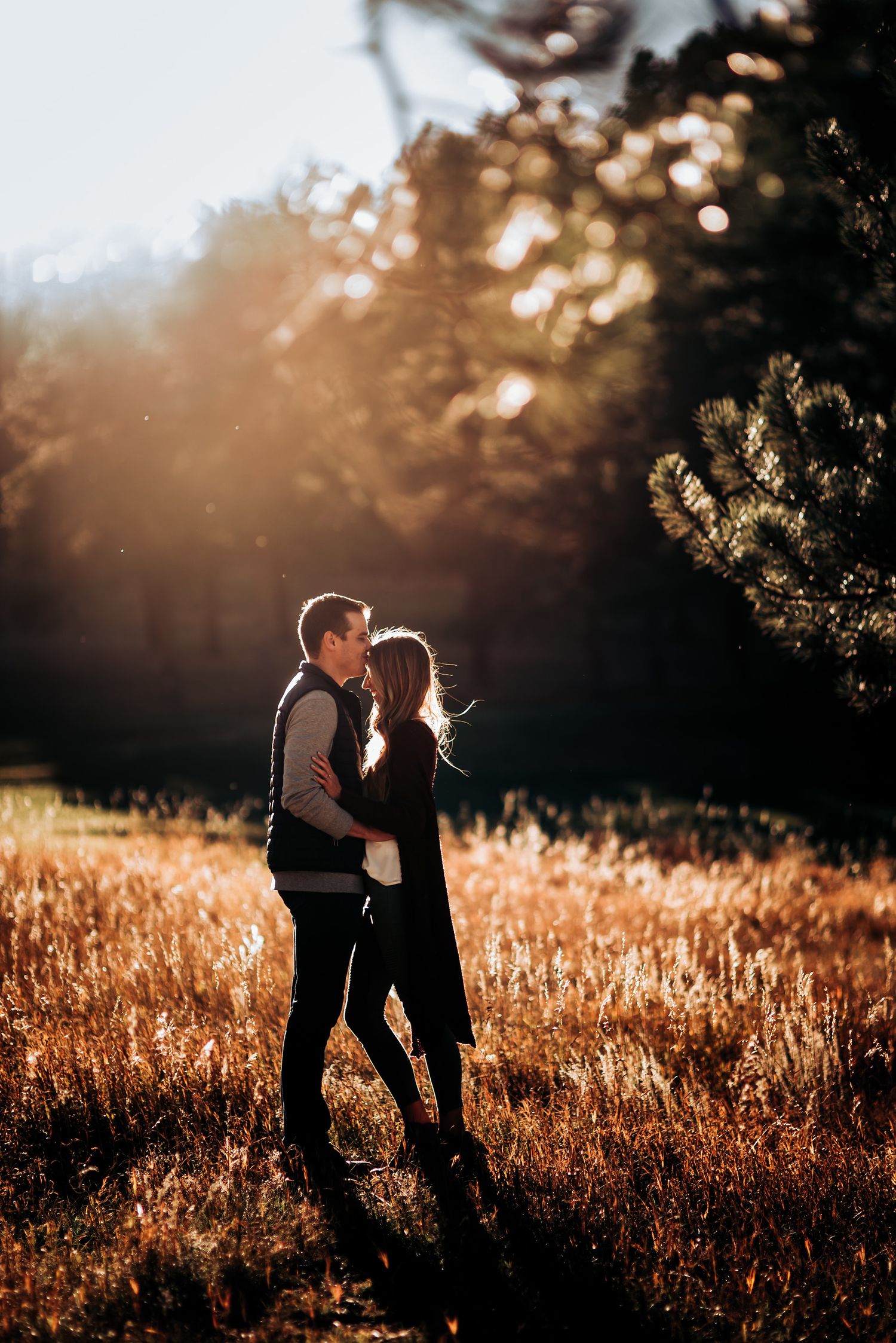 Two silhouettes stand together in a sunlit field during golden hour with trees in the background.