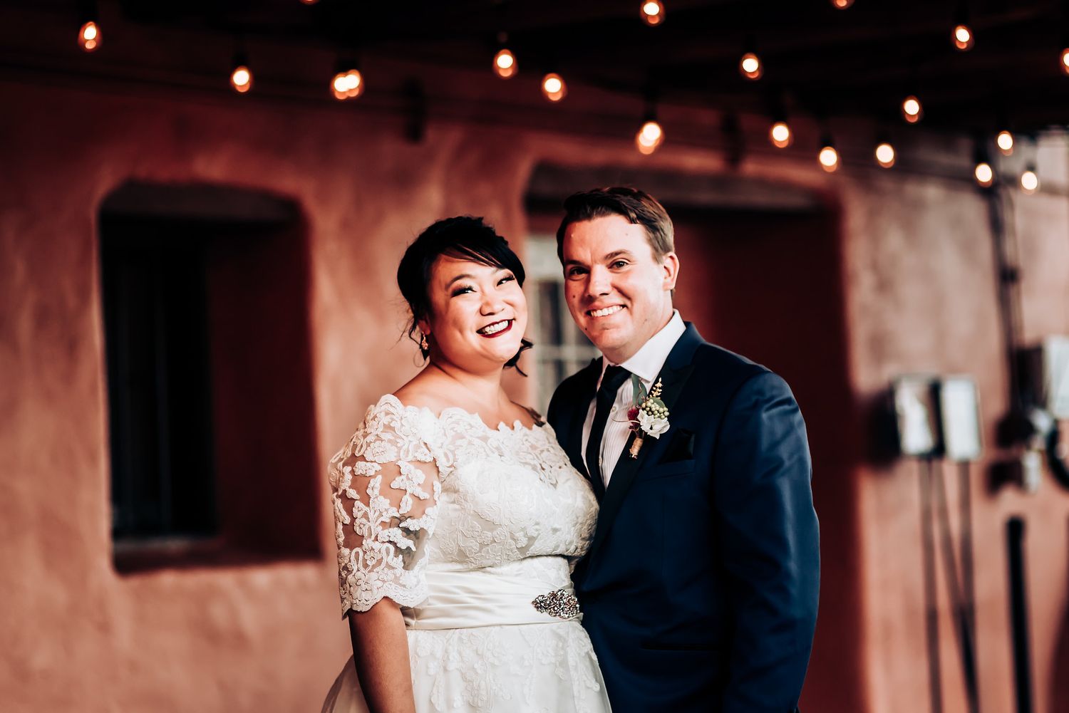 Evening wedding portrait under string lights with warm adobe walls in background.