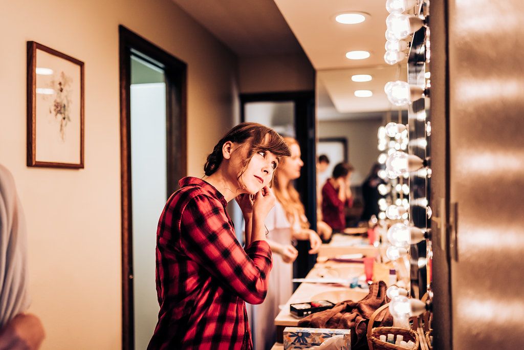 People getting ready at a makeup station with bright vanity lights and a red plaid shirt visible.