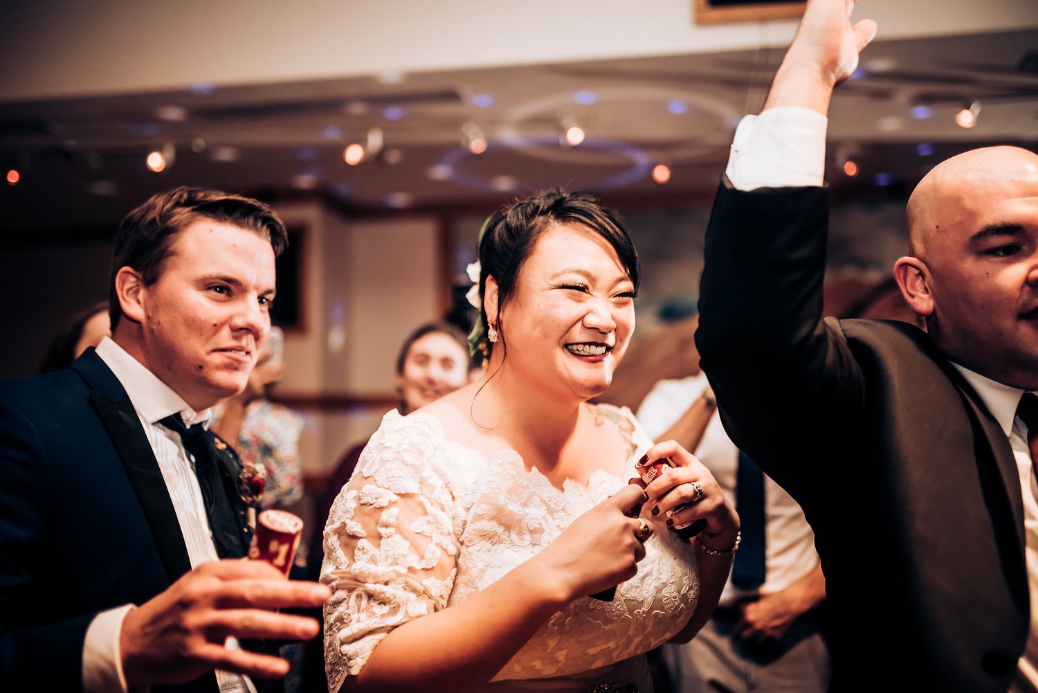 Wedding party celebrating with drinks and raised arms under indoor lighting.