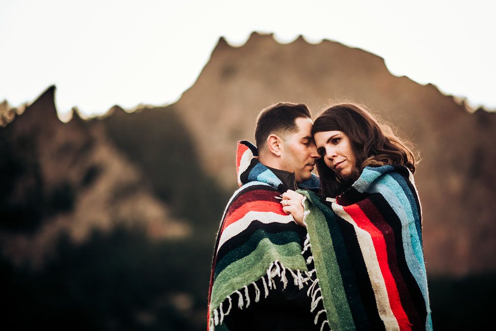 A romantic mountain portrait of a couple wrapped in a striped blanket against a dramatic backdrop.