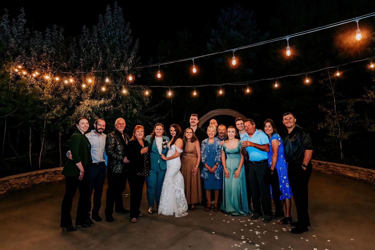 Wedding party and family group photo outdoors at night under warm string lights.
